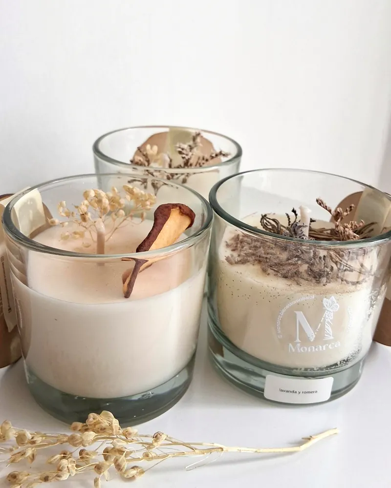 Tres velas de vidrio con flores secas, pera deshidratada y lavanda, junto a un ramito seco, sobre una mesa blanca