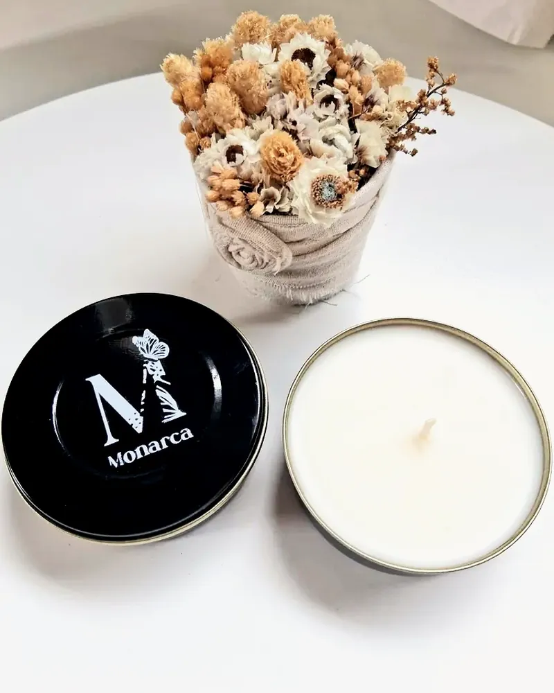 Vela en lata con tapa negra con el logo de Monarca, junto a un ramito de flores secas