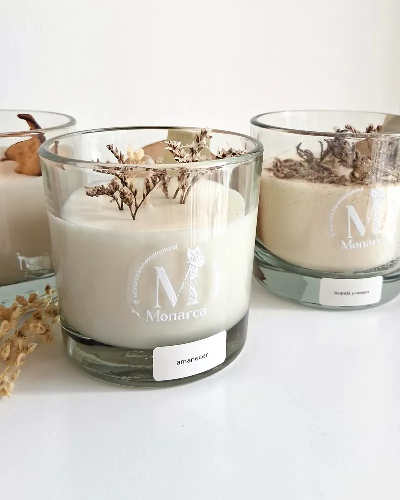 Velas con magia: vela en vaso de vidrio con flores secas y el logo de Monarca, junto a un ramito de flores secas